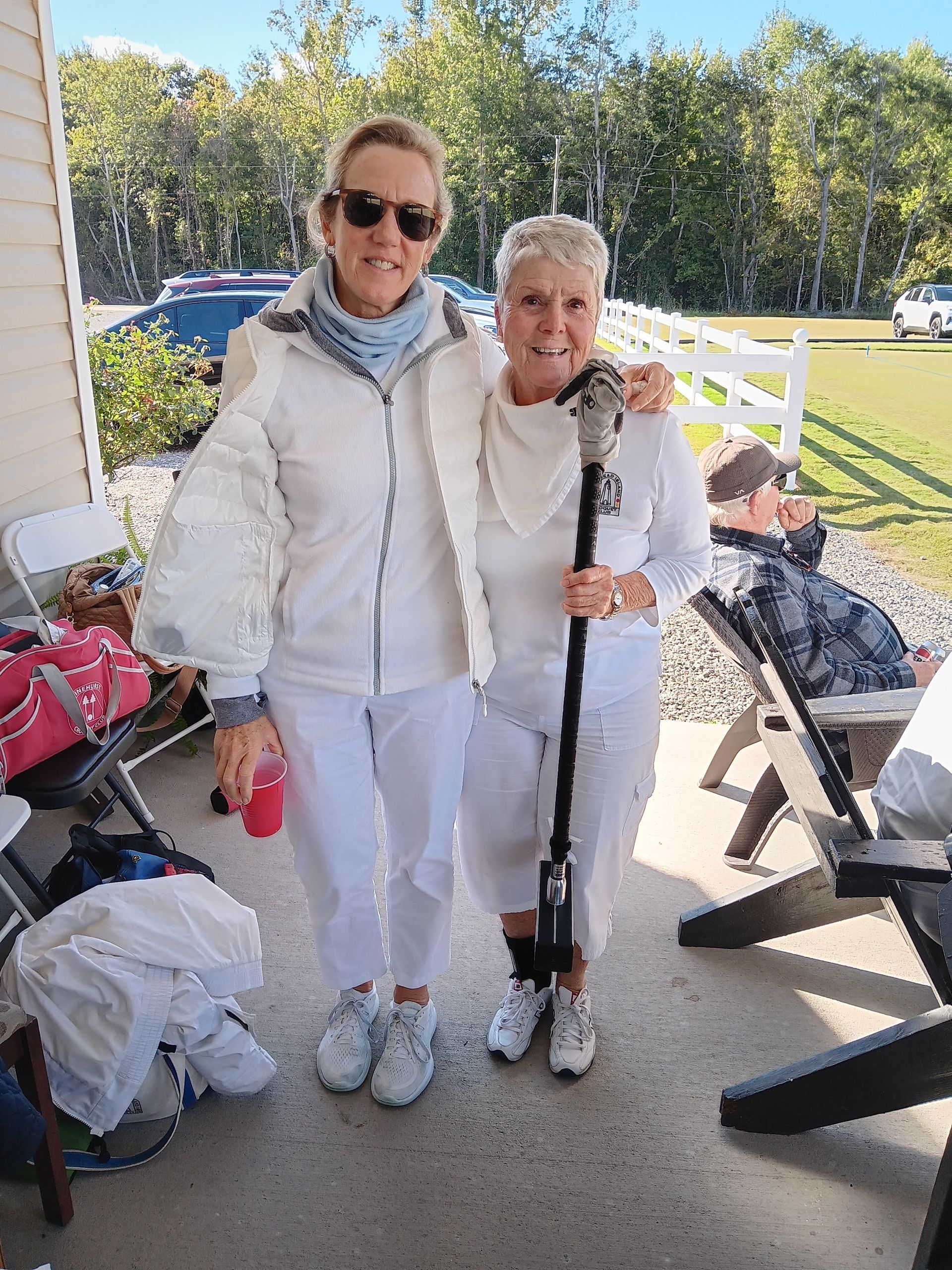 Two women in white outfits, one with a cane, standing on a porch. Sunny outdoor setting.