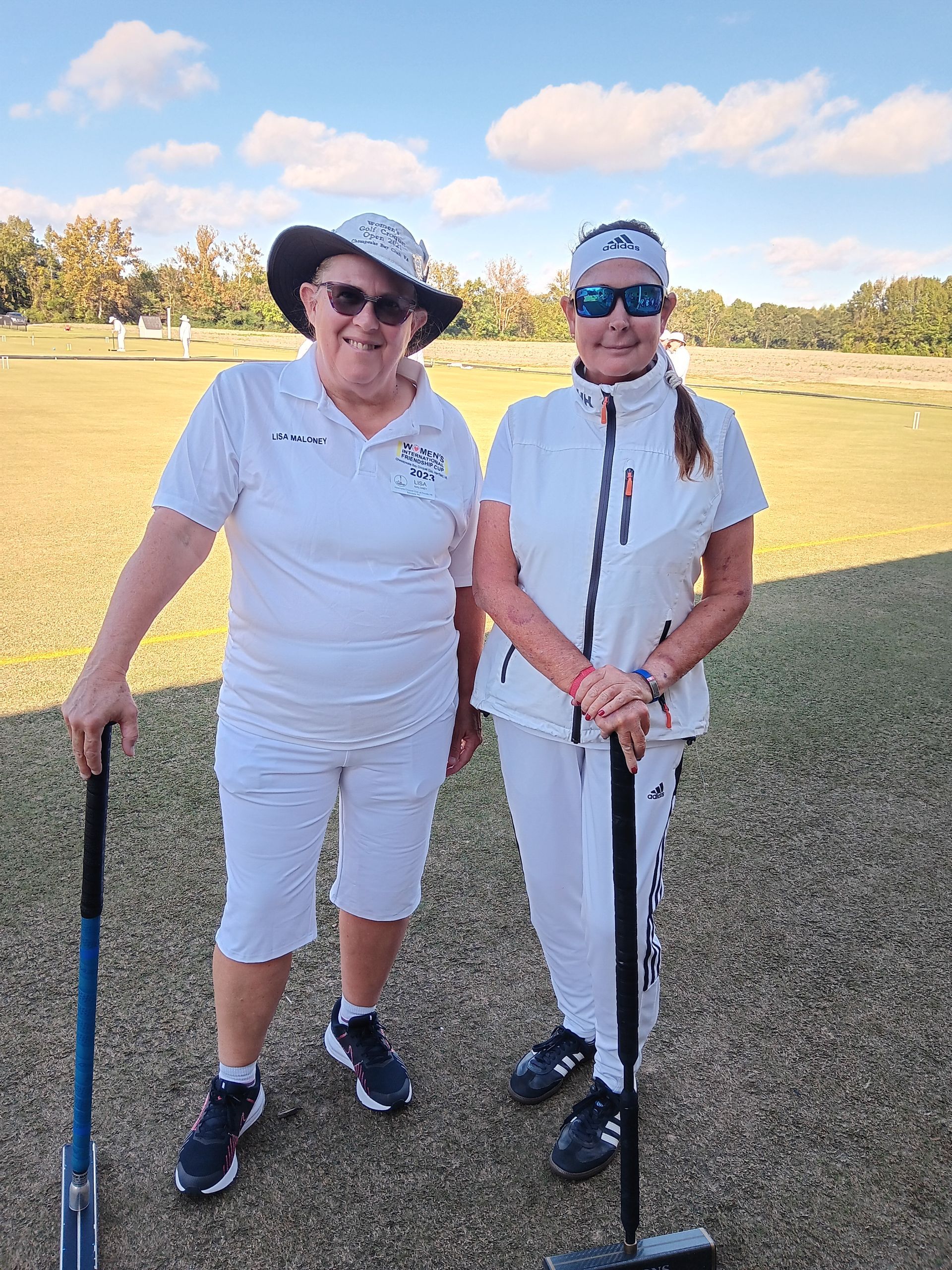 Two women in white sport attire stand on a grassy field, holding mallets. Blue sky and trees are in the background.