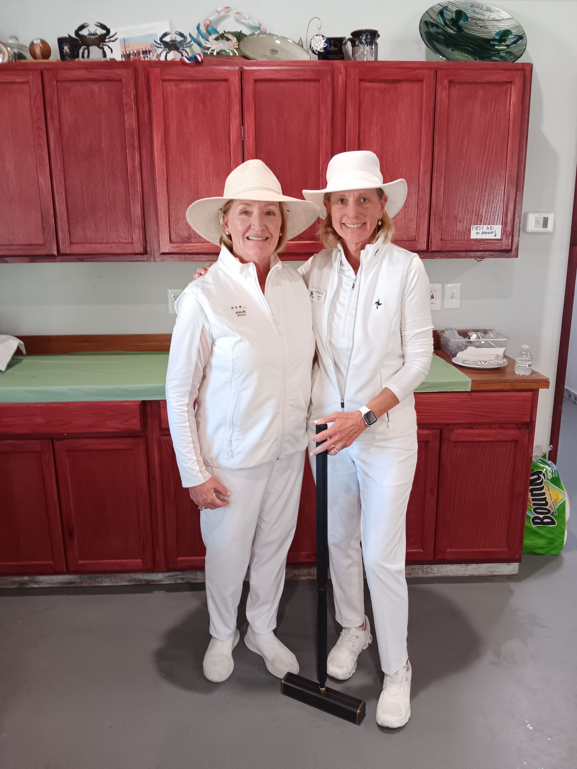 Two women in white outfits and hats pose with a croquet mallet in a room with red cabinets.