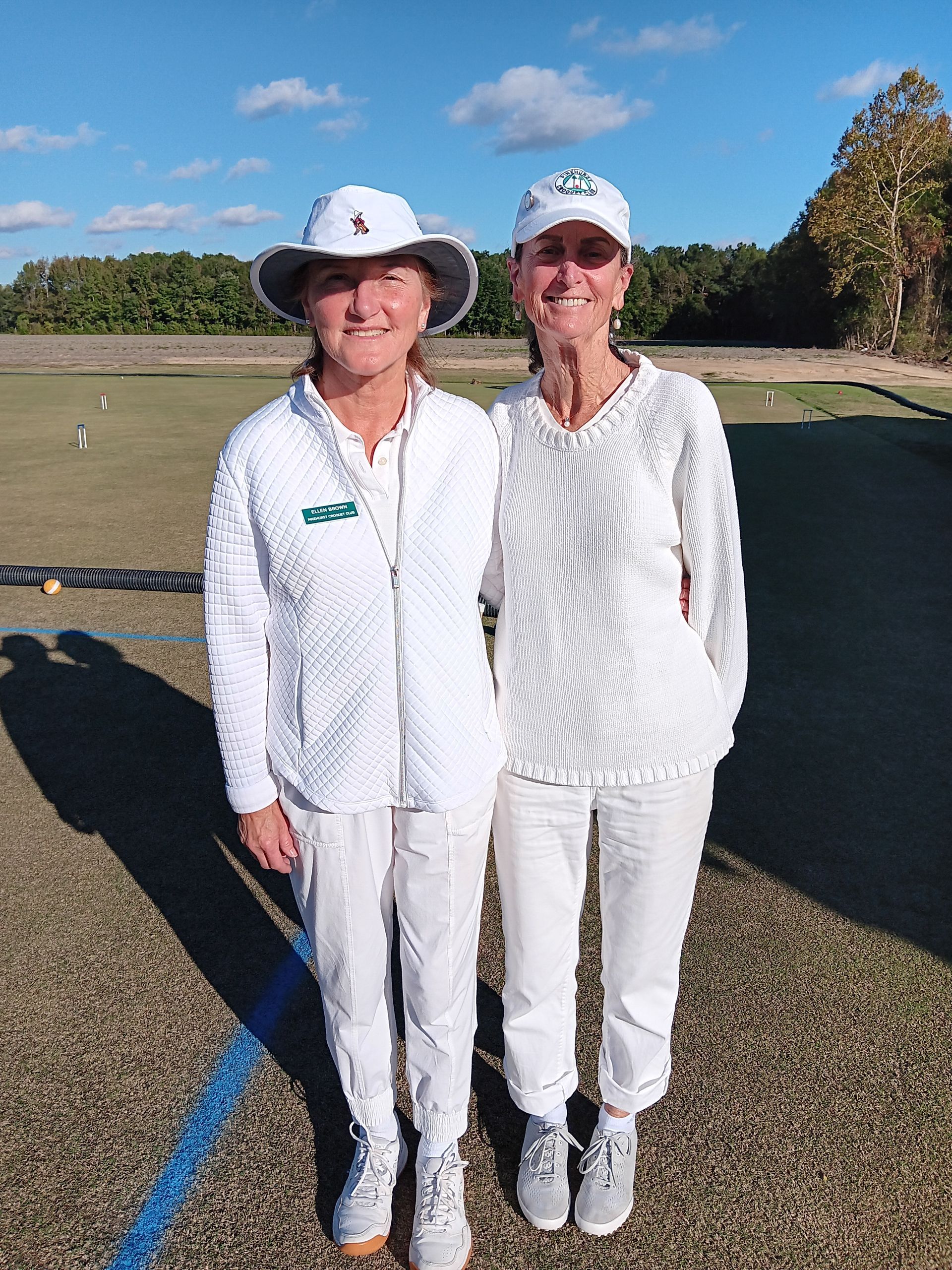 Two women in white outfits standing on a grass field, posing for a photo.
