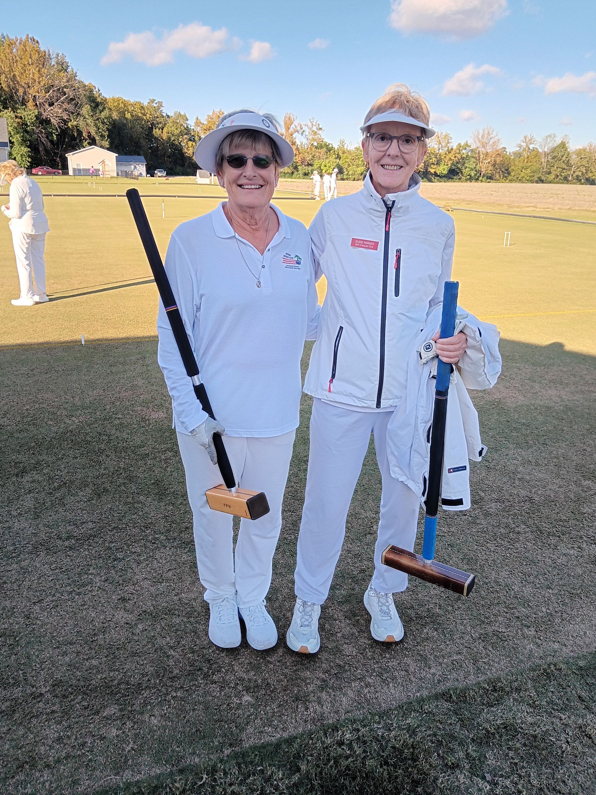Two women in white outfits on a croquet field, each holding a mallet.