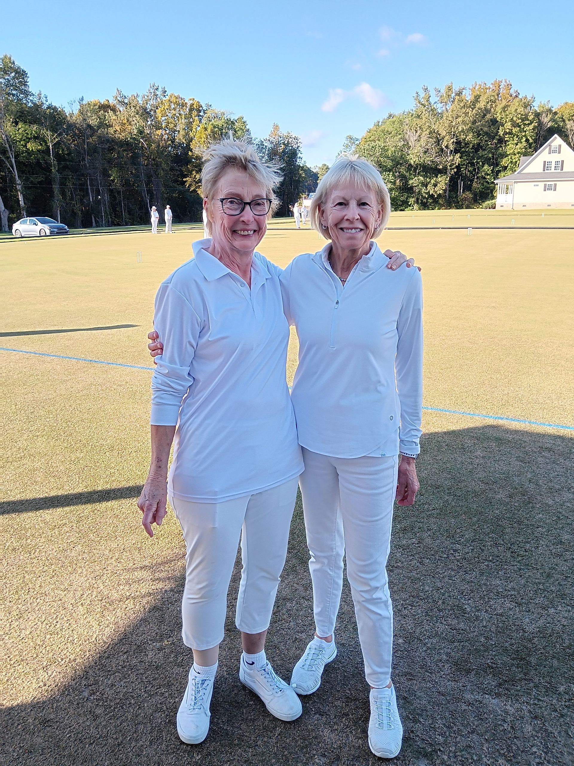 Two women in white outfits, standing on a grassy field, arms around each other, smiling.