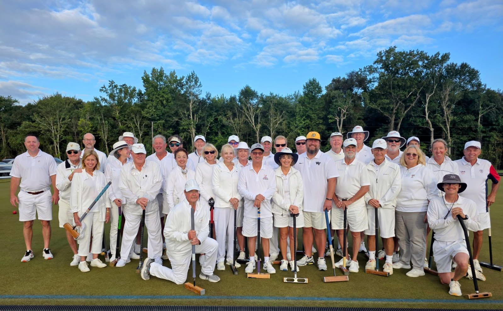 Group of people in white attire, holding croquet mallets on a green field, posing outdoors.