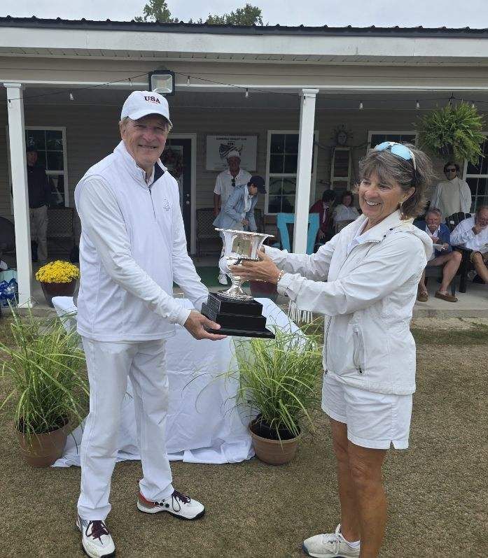 Man in white handing a trophy to a woman in white, outdoors near a building.