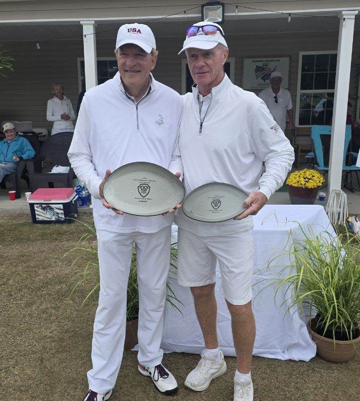 Two men in white golf attire holding awards on a course.