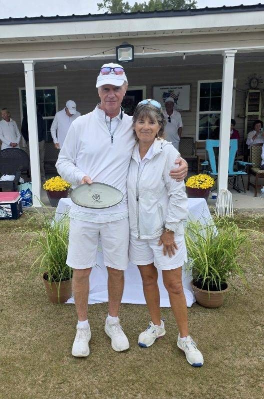 Man and woman in white, embracing. Man holds a trophy, standing in front of a building and greenery.
