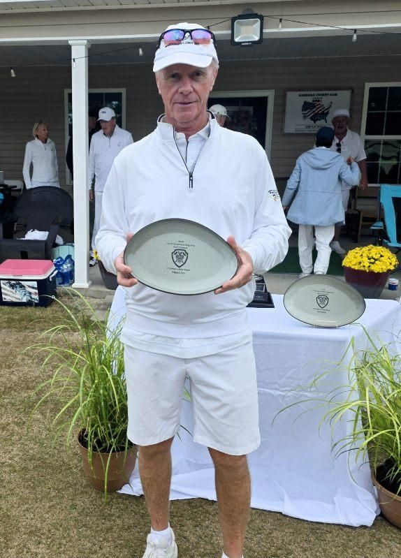 Man in white golf attire holds a trophy outdoors; other people, plants, and tables in background.
