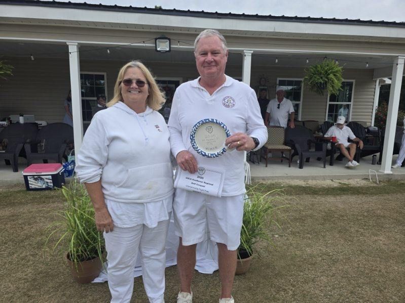 Man and woman in white outfits holding an award, standing in front of a building with people.