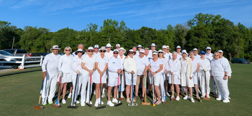 A large group of people in white attire, holding croquet mallets, pose on a green lawn. Blue sky, trees.