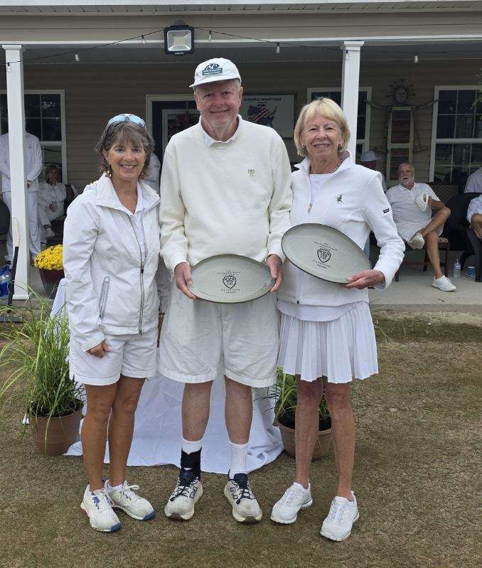 Three people in white attire holding plates, posing on a lawn in front of a building.