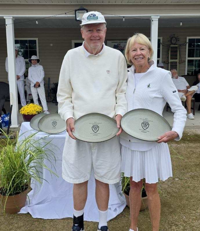 Two people hold plates; standing outside near a table with more plates, other people, and foliage.