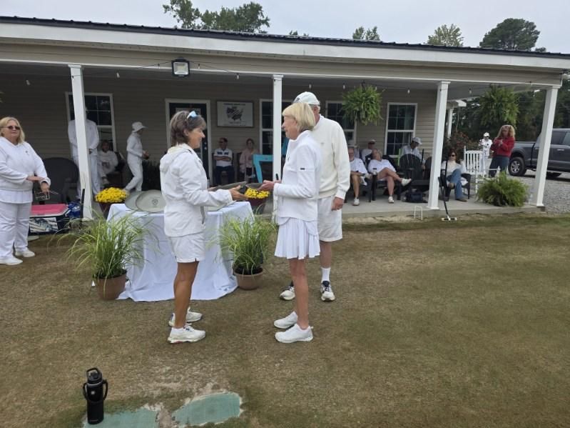 Two people in white attire are presented with an award on a lawn near a building.