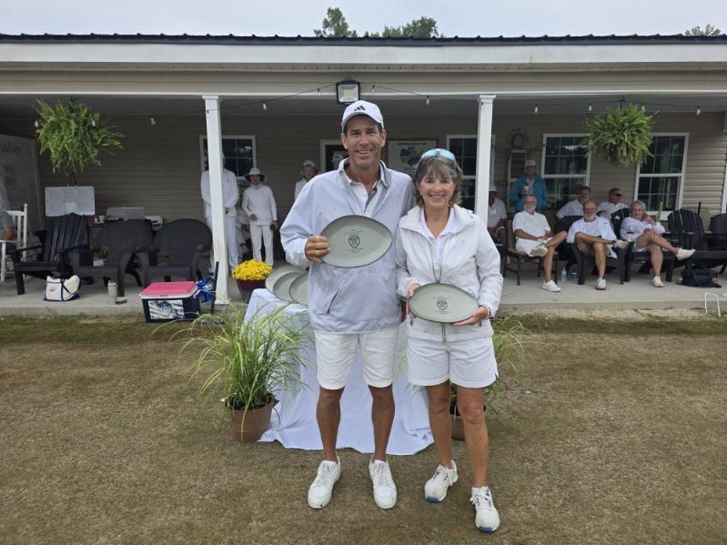 Man and woman in white holding plates, smiling, posed in front of a building with people seated on a lawn.