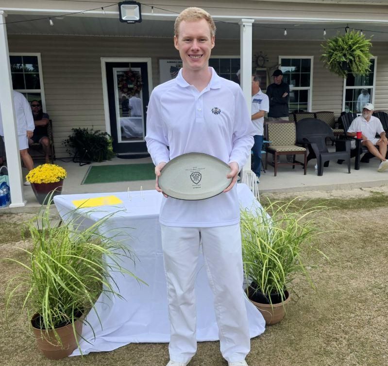 Man holding trophy at outdoor event. White shirt and pants, green grass, building in background.