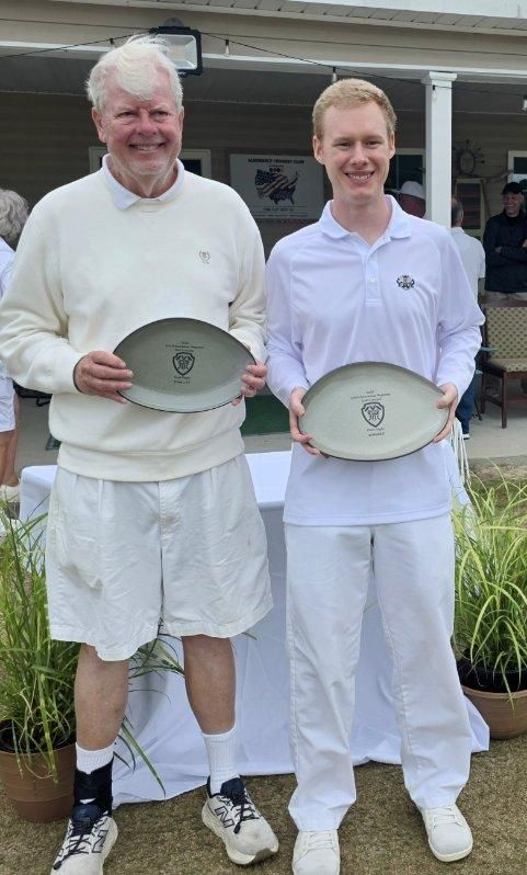 Two people holding plates outdoors. One in shorts and sweater, the other in pants and shirt, both white.