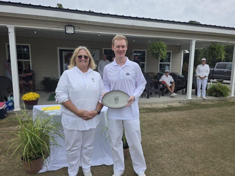 Two people in white outfits stand outside, holding a trophy. Building and spectators in background.