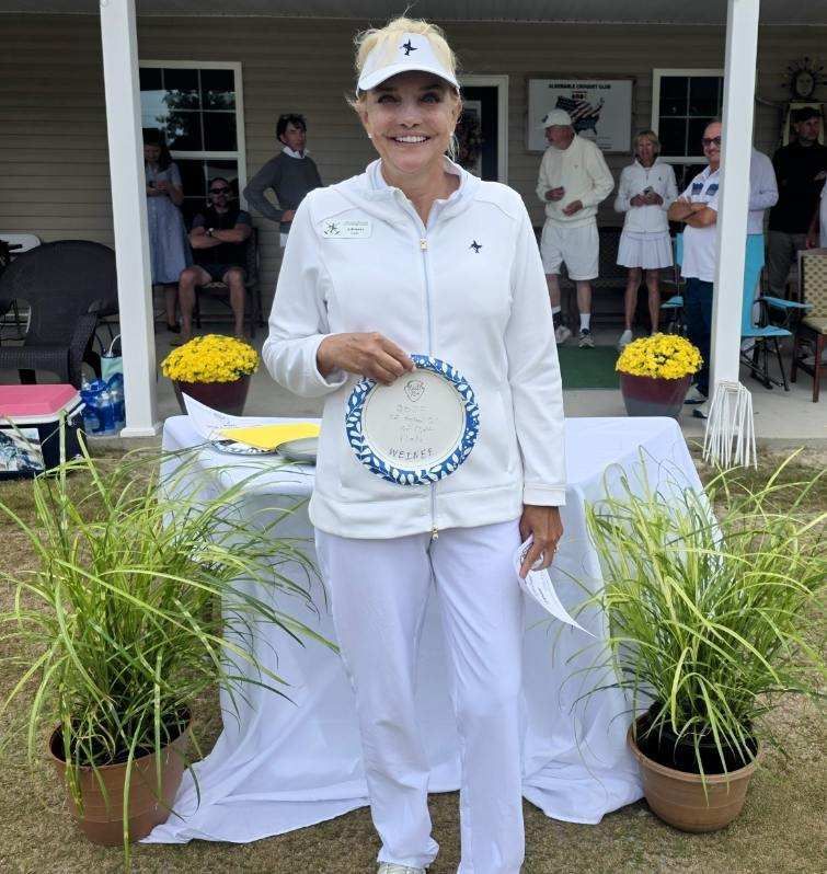 Woman in white golf attire holding award, smiles, standing outdoors near flowers.
