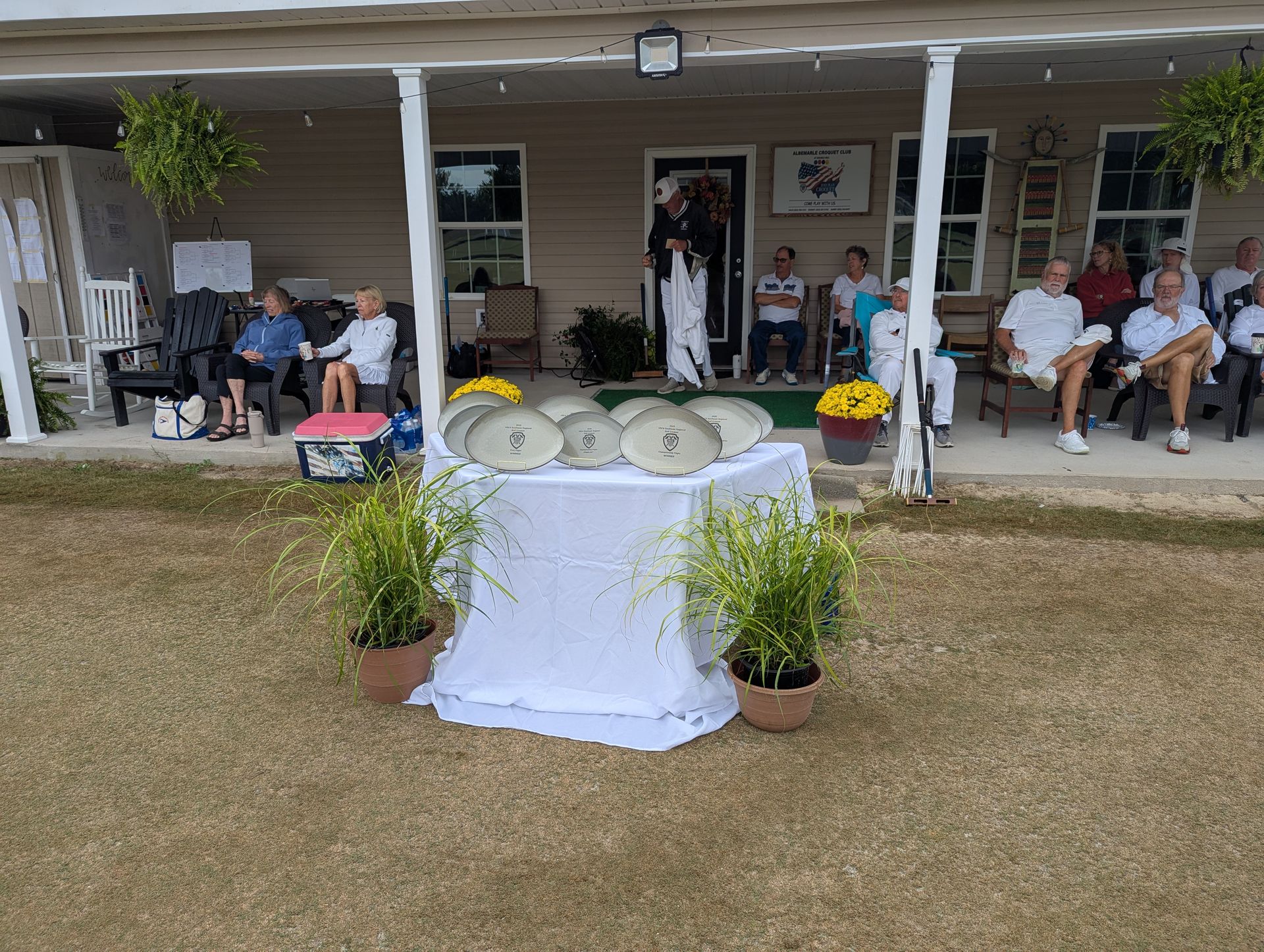 People seated on a porch, observing a table with plates and plants outdoors.