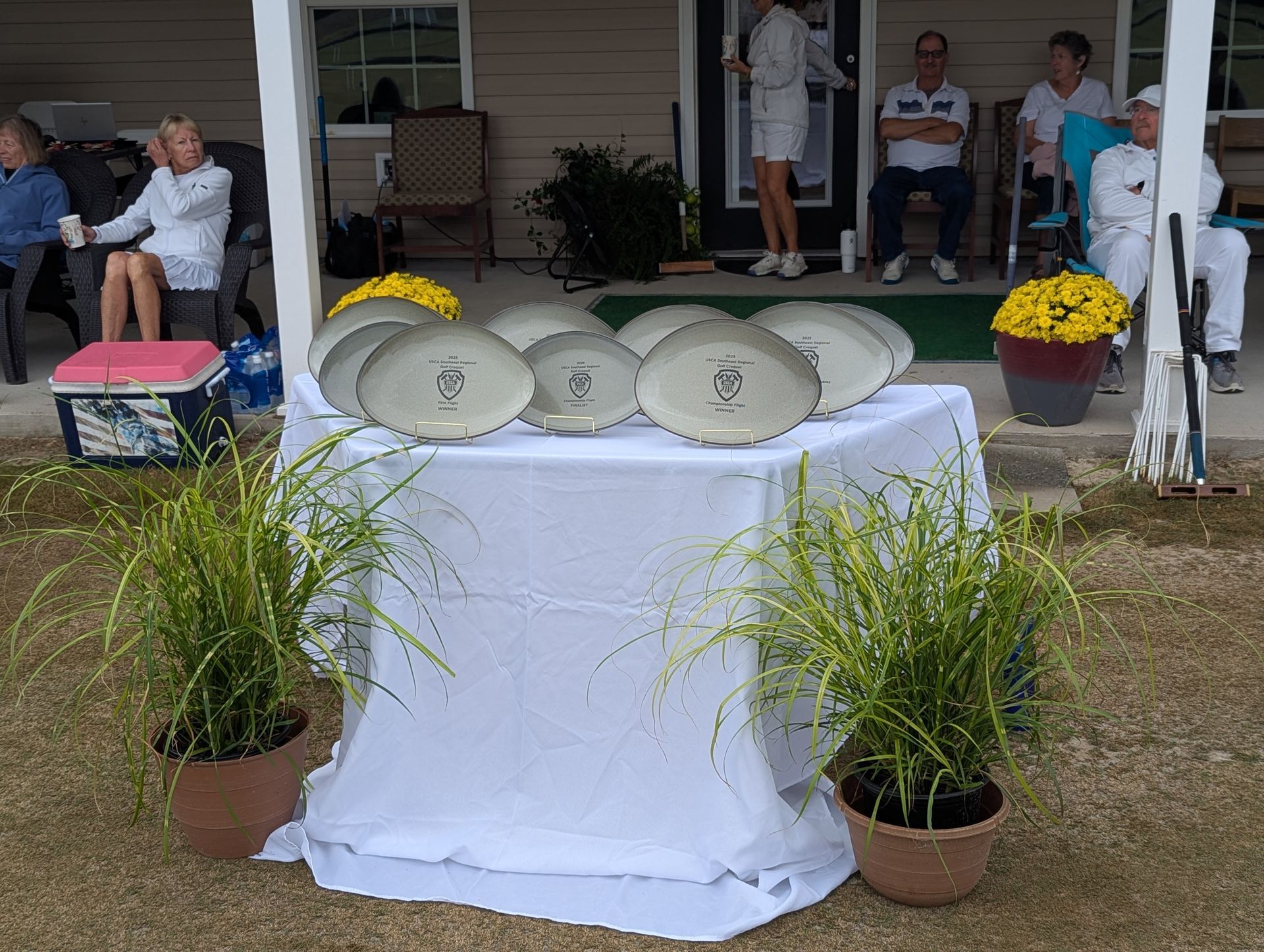 Oval plates on a table outdoors, with people watching. Potted plants flank the table.