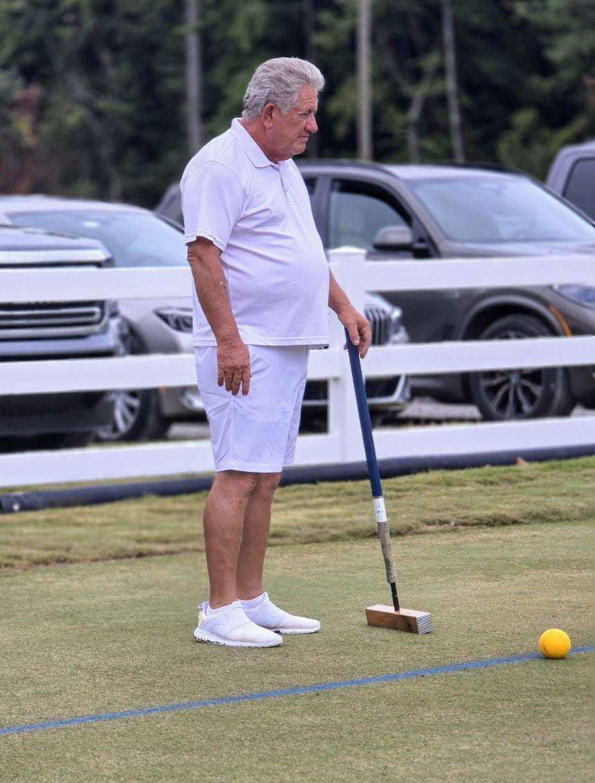 Man in white playing croquet, holding mallet, next to yellow ball on a grass lawn. Cars are in the background.