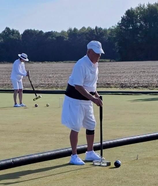 Two people in white playing croquet on a green field. One bends over, focused, while the other prepares to swing.