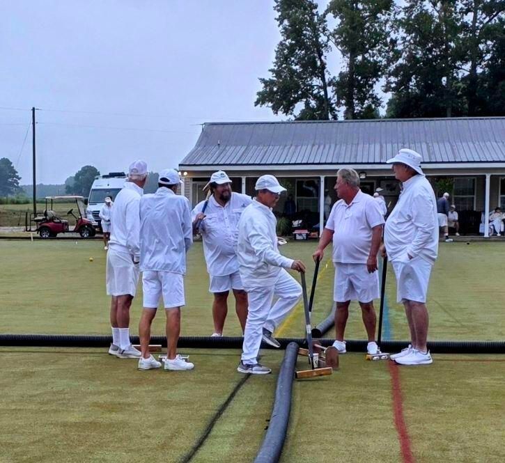 Group of men in white uniforms on a green lawn, near a building. They appear to be playing a lawn game, looking at equipment.