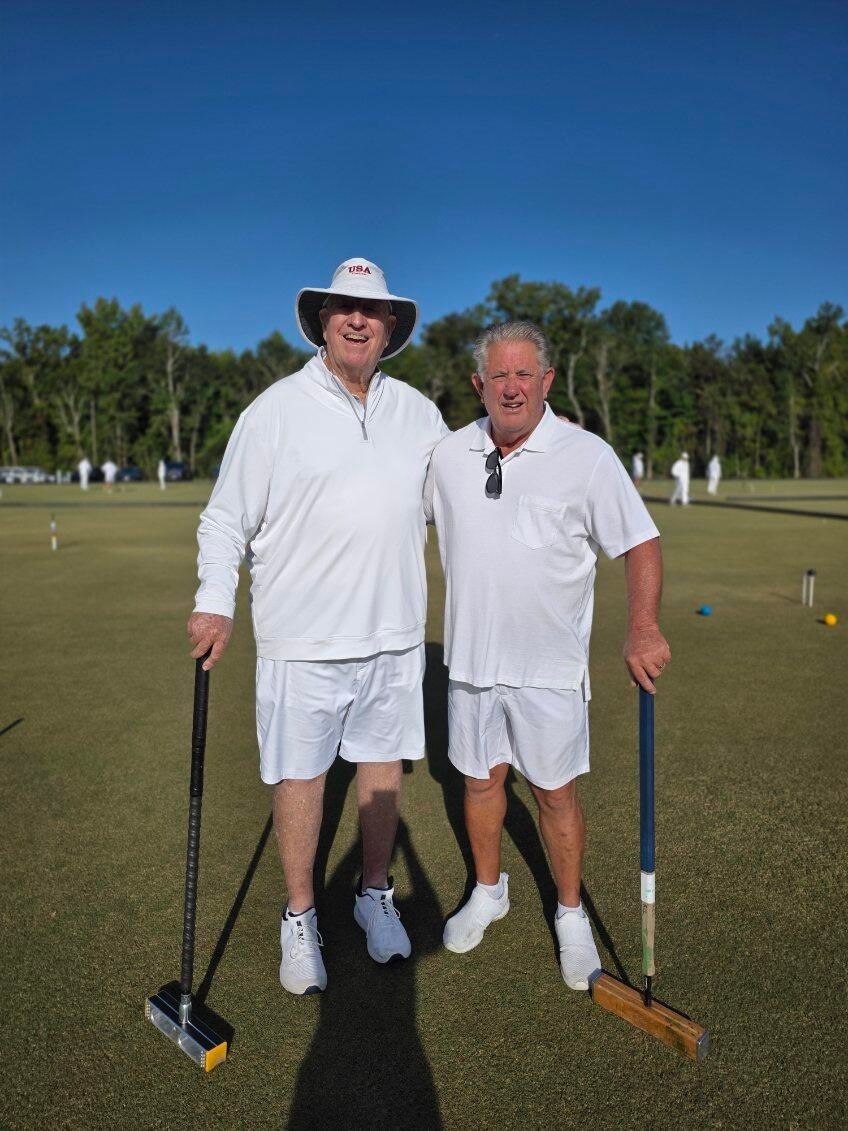 Two men in white clothing, holding mallets, pose on a croquet lawn. Sunny day, trees in background.
