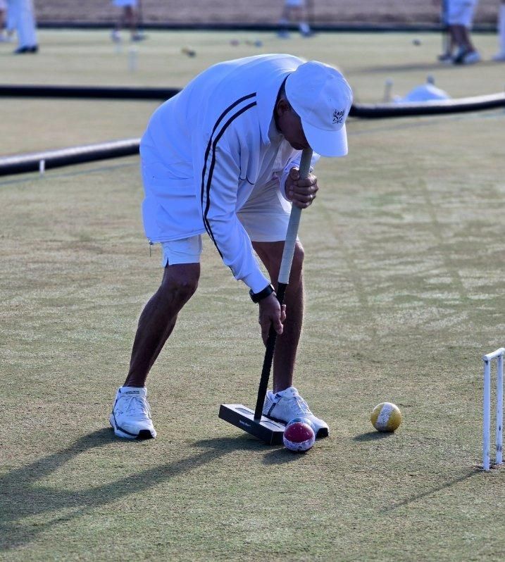 Man in white clothing playing croquet, bending over to strike a ball with a mallet on a green lawn.