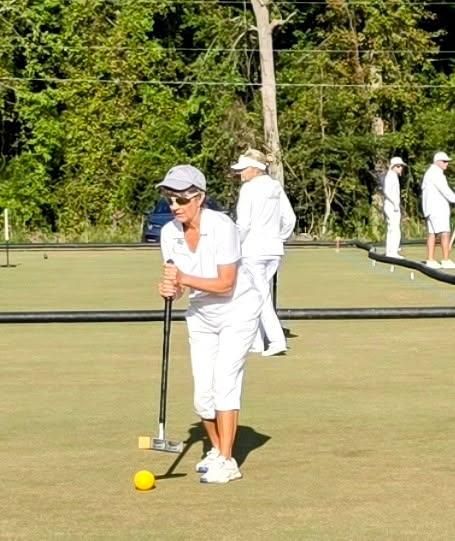 Woman in white outfit aiming at yellow croquet ball on green lawn.