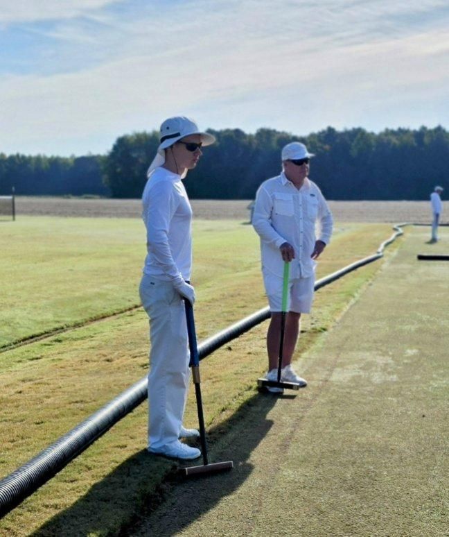 Two people in white attire on a croquet lawn, holding mallets. Sunny day.