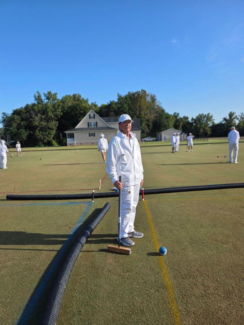 Man in white croquet attire on a green field, holding mallet, with other players and a house in the background.