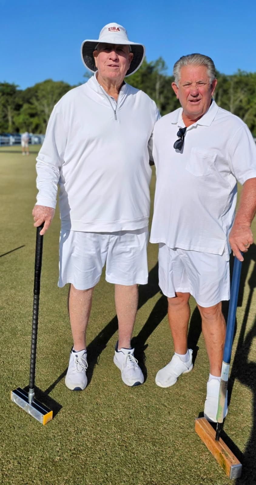 Two men in white outfits with croquet mallets on a green lawn.