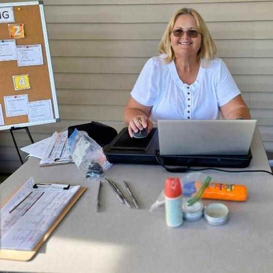 Woman at a table with laptop, supplies, and clipboard, smiling. Corkboard in the background.