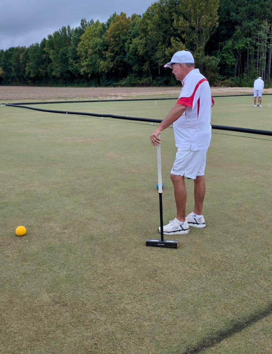 Man in white playing croquet on a green lawn; holds mallet near yellow ball.
