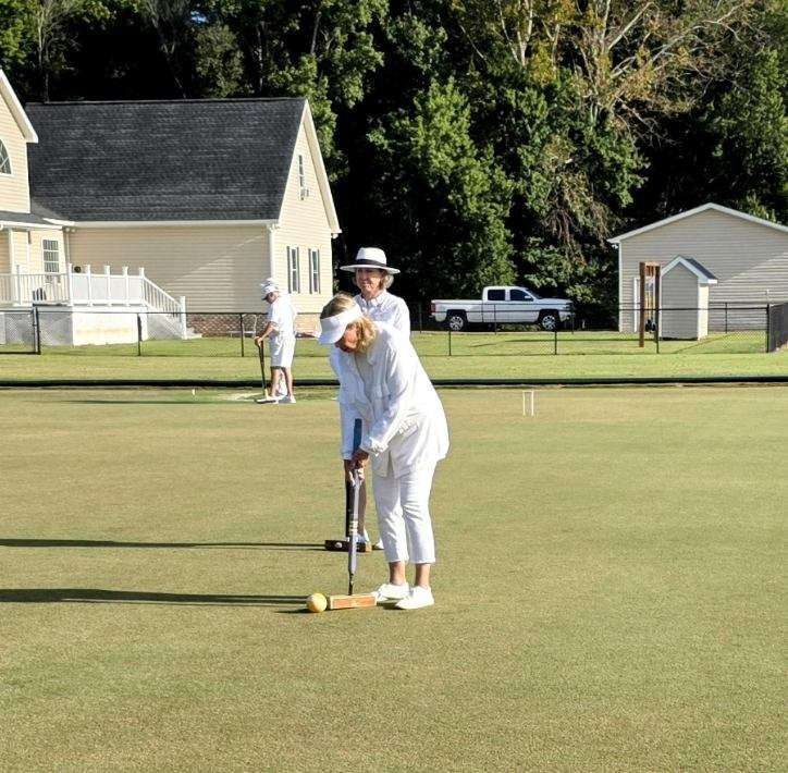 People in white attire playing croquet on a green lawn with buildings in the background.