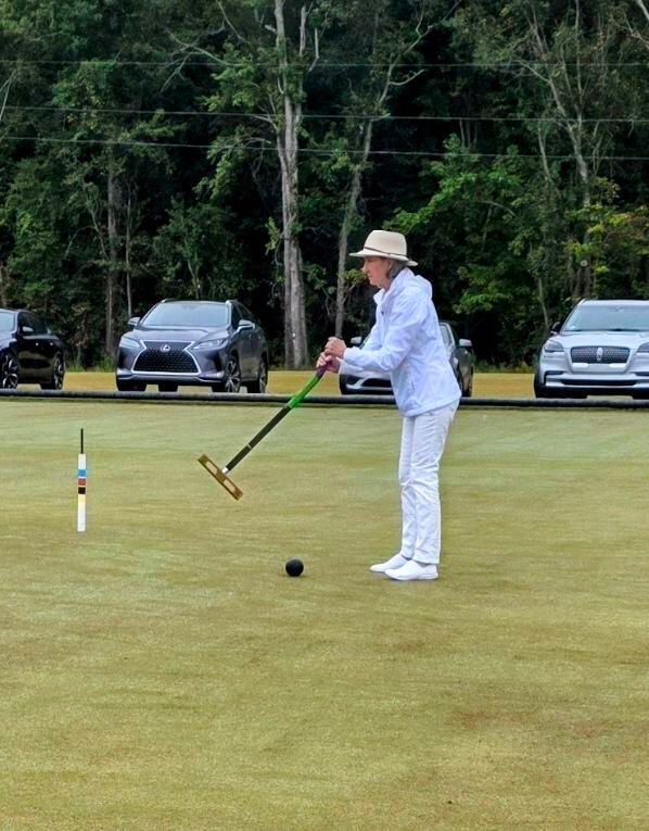 Person playing croquet on a green lawn, holding mallet, near a black ball and stakes. Cars in the background.