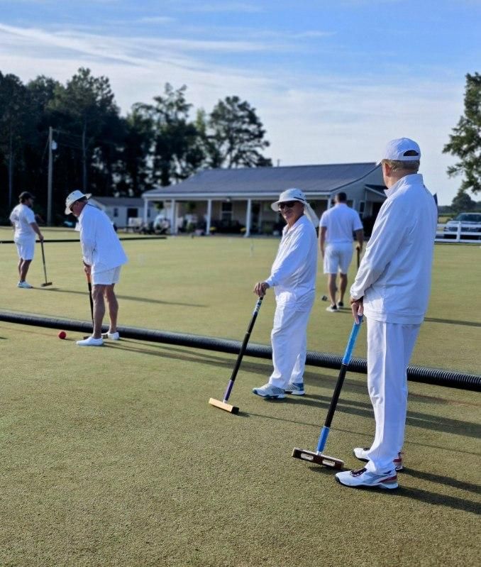 People playing croquet on a green lawn, wearing white clothing. A clubhouse is in the background.
