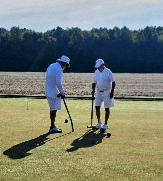 Two men in white outfits play croquet on a green lawn, with a field and trees in the background.