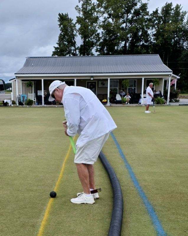 Man in white clothing playing lawn bowls on a green field with a clubhouse in the background.