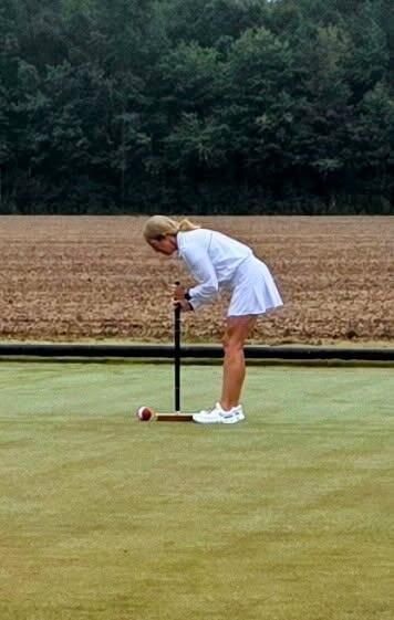 Woman in white plays croquet on a green lawn, bent over a ball with a mallet.