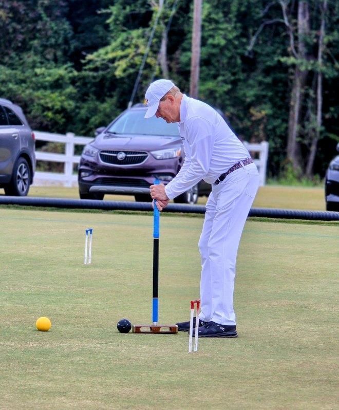 Man in white playing croquet on a green lawn, aiming toward a hoop.