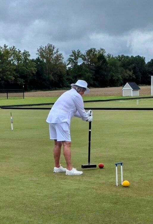 Person in white outfit plays croquet on a green lawn with mallets and balls near white hoops. Overcast sky in background.