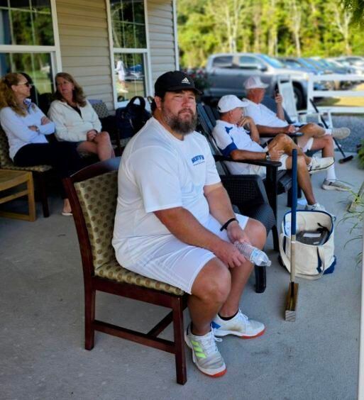 A group of people sitting on chairs outdoors. One man in white tennis attire looks at the camera.