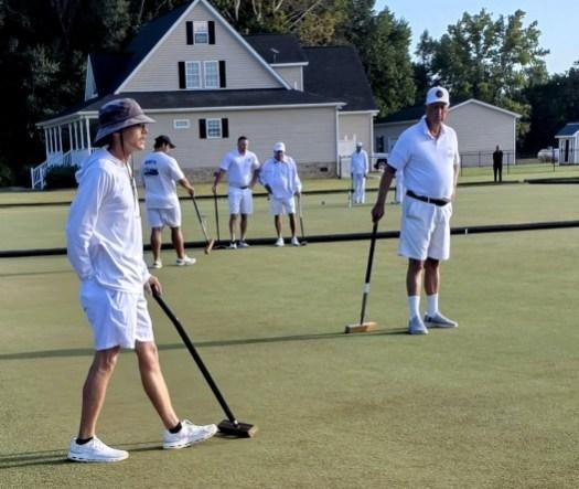 People playing croquet on a green lawn. Players wear white and use mallets. House in background.