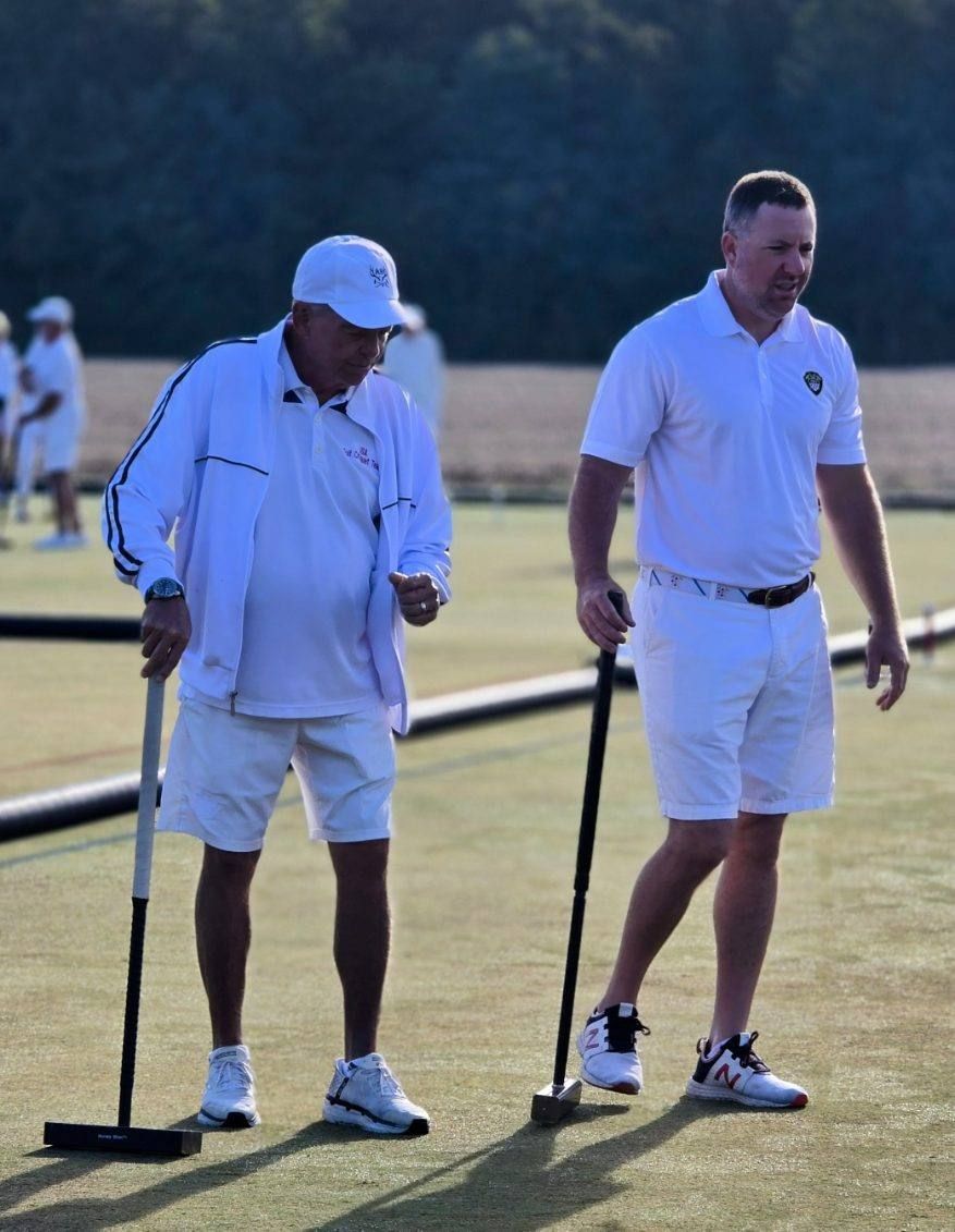 Two men in white attire playing croquet on a green field. One man is looking down, the other looks forward.