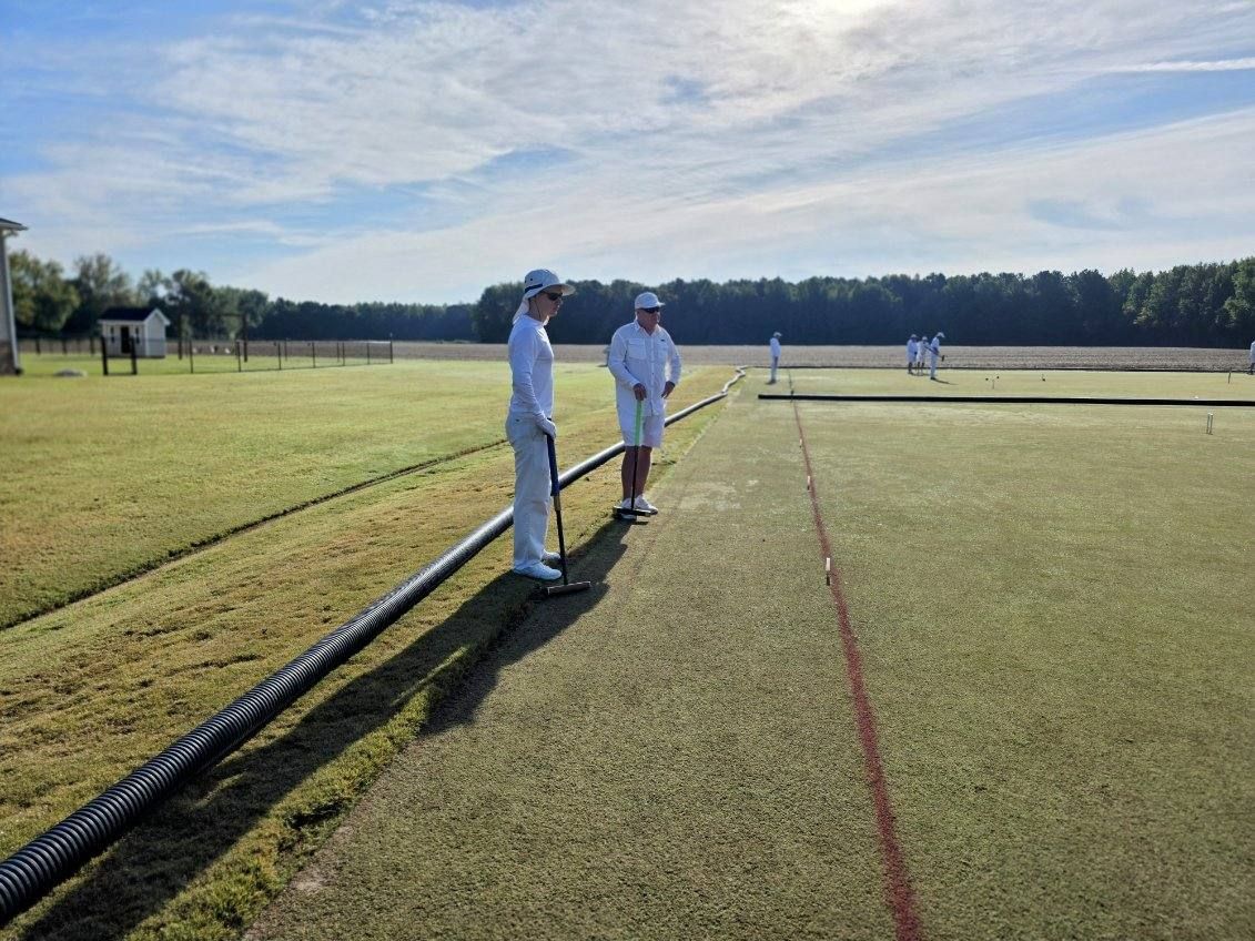 Two people in white outfits stand on a croquet lawn, preparing to play.