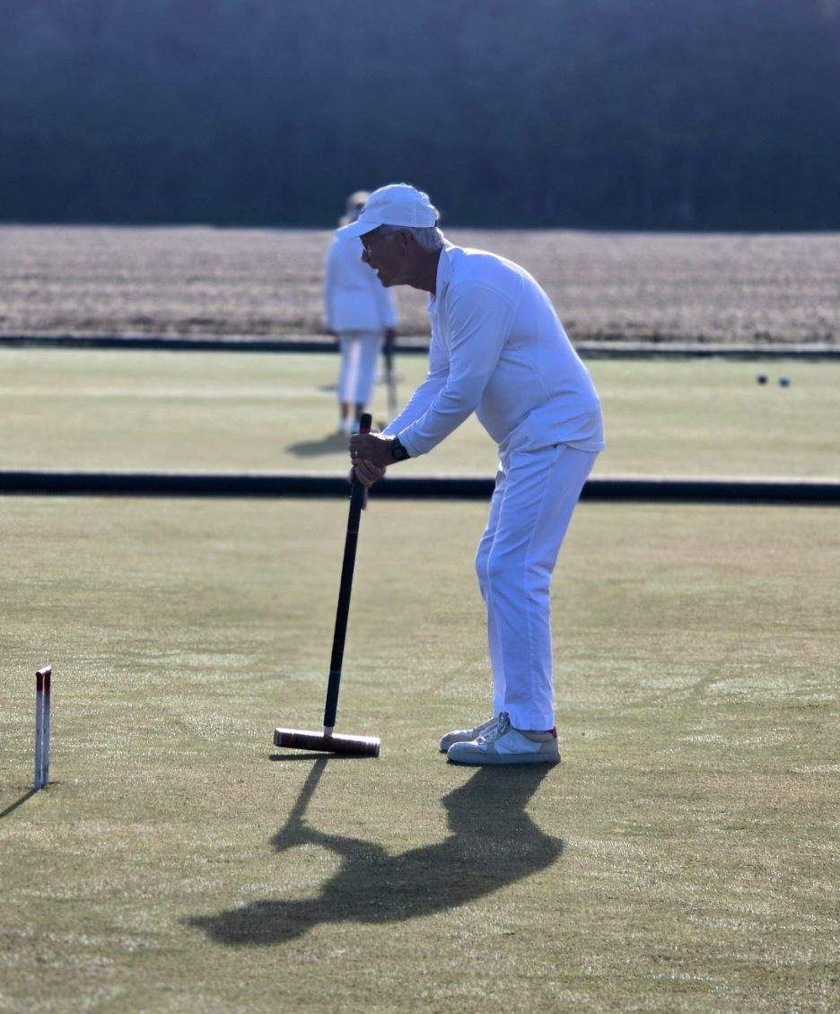 Man in white outfit playing croquet on a green lawn, holding mallet, with shadows.