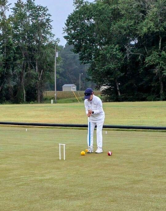 Person playing croquet on a green lawn, holding a mallet. A yellow ball is near a hoop.
