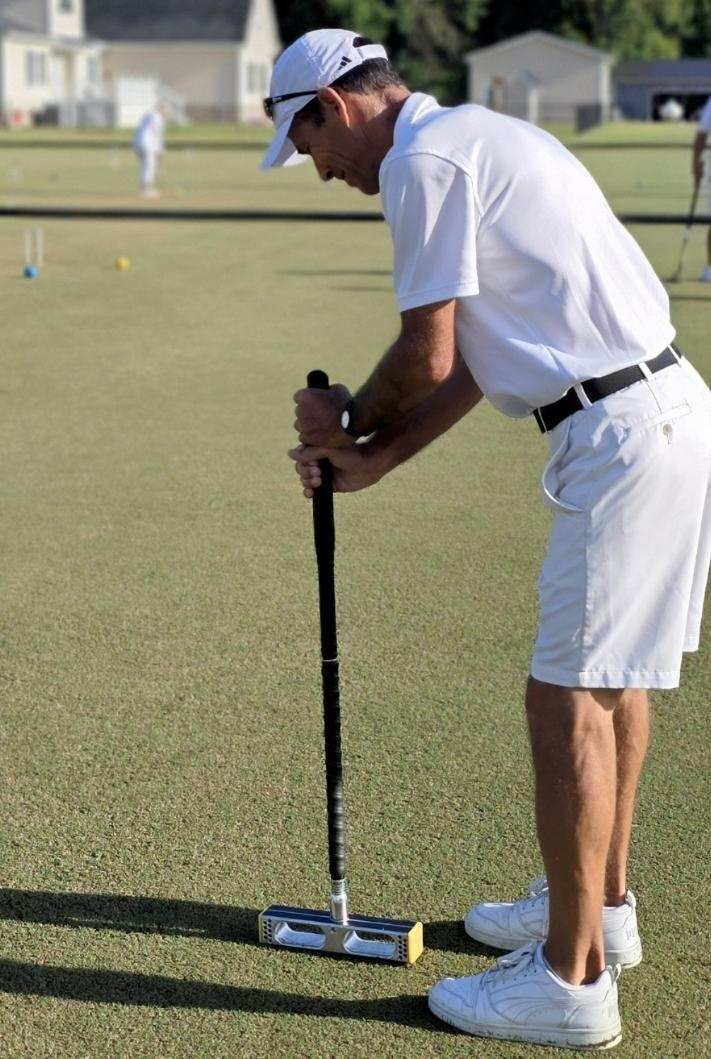 Man in white clothing playing croquet, bending over mallet on green lawn.