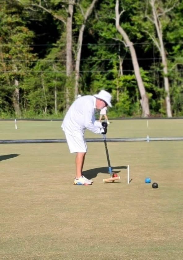 A person in white playing croquet on a green lawn, aiming at a ball with mallet, wearing a white hat.